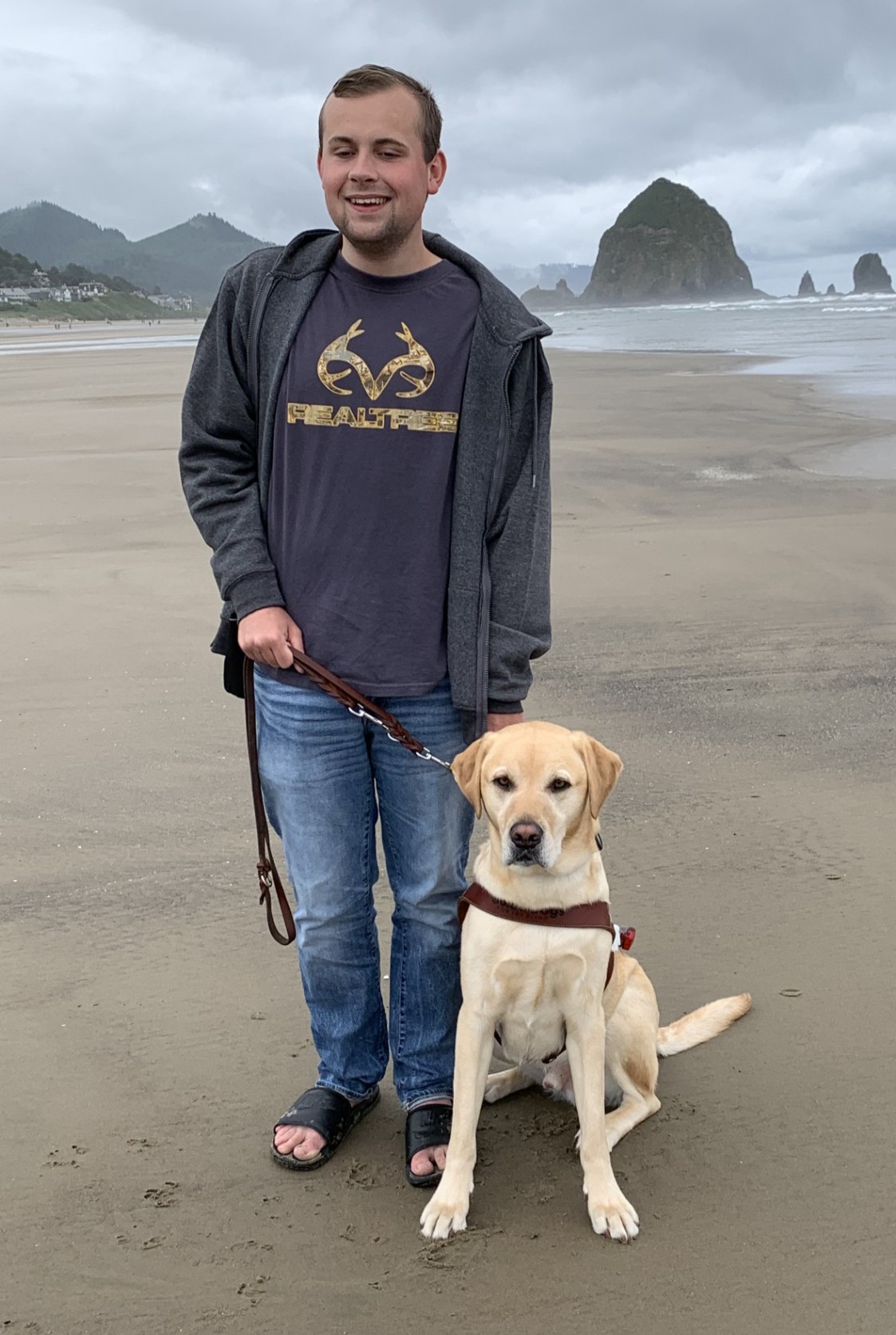Ethan stands on a sandy beach with his yellow Lab guide dog, Ginsey, sitting by his side. Large rock formations and the ocean are visible in the background under a cloudy sky.
